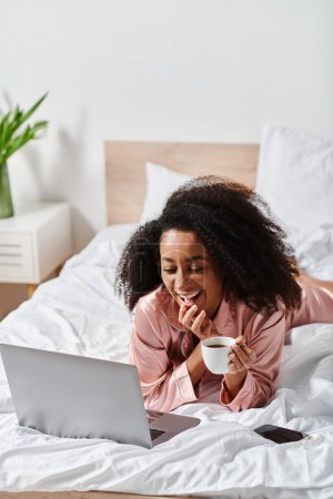 A curly African American woman in pajamas peacefully lays on a bed, enjoying a laptop and a cup of coffee in her bedroom in the morning.