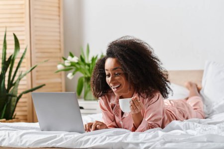 Curly African American woman in pajamas peacefully laying on bed, working on laptop in cozy bedroom during morning time.