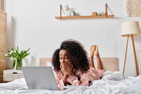 Téléchargez les photos : Femme afro-américaine bouclée en pyjama couchée au lit, absorbée par l'utilisation d'un ordinateur portable. - en image libre de droit