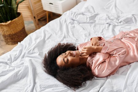 Photo for A curly African American woman in pajamas relaxes on a white bed in a serene morning setting. - Royalty Free Image