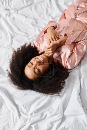 A curly African American woman in pajamas peacefully rests on a white bed in the morning light.