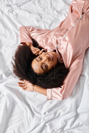 Una mujer afroamericana rizada en pijama tendida pacíficamente sobre una cama blanca en un dormitorio durante la mañana.