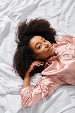 A curly African American woman in pajamas peacefully lays on top of a white sheet in her bedroom during morning time.