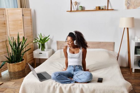 Téléchargez les photos : A curly African American woman in a tank top sits on a bed, engrossed in her laptop. - en image libre de droit