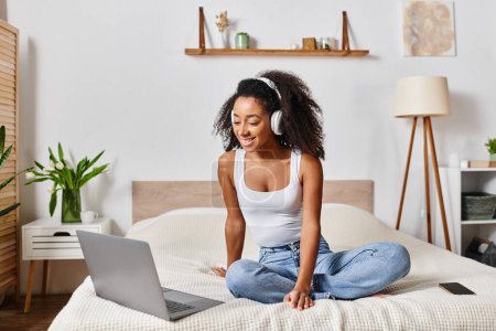 Eine lockige Afroamerikanerin im Tank-Top sitzt auf einem Bett, tief konzentriert auf ihrem Laptop in einem modernen Schlafzimmer.