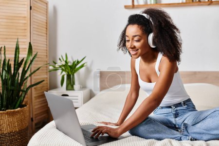 Mujer afroamericana rizada en una camiseta sin mangas, sentada en una cama, intensamente enfocada en usar una computadora portátil en un dormitorio moderno.