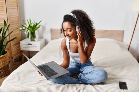 Foto de Rizado mujer afroamericana en camiseta sin mangas se sienta en la cama con ordenador portátil en el elegante dormitorio moderno. - Imagen libre de derechos