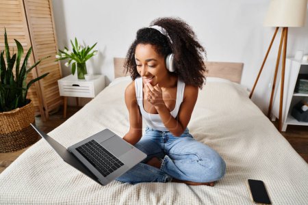 A curly African American woman in a tank top is sitting on a bed, using a laptop and wearing headphones in a modern bedroom.