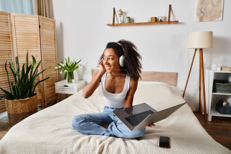 Mujer afroamericana rizada en una camiseta sin mangas sentada en una cama, usando una computadora portátil y usando auriculares en un dormitorio moderno.