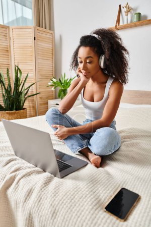 Téléchargez les photos : A curly African American woman sits on a bed, focused on her laptop screen in a modern bedroom. - en image libre de droit