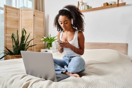 Lockige Afroamerikanerin in einem modernen Schlafzimmer auf dem Bett sitzend, in einen Laptop vertieft.