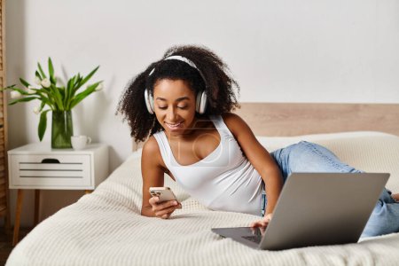Foto de Mujer afroamericana rizada en una camiseta sin mangas tumbada en una cama, absorbida por el uso de una computadora portátil y con auriculares. - Imagen libre de derechos