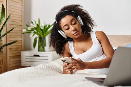 Una mujer afroamericana rizada en una camiseta sin mangas se relaja en la cama, escuchando música en su teléfono en un dormitorio moderno.