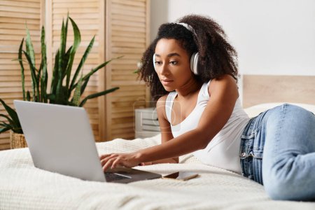 A curly African American woman in a tank top lies on a modern bed, intensely focused on using a laptop computer.