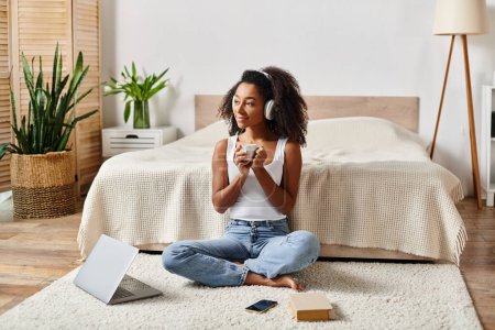 Téléchargez les photos : Femme afro-américaine bouclée assise sur le sol devant un ordinateur portable dans une chambre moderne, travaillant ou étudiant. - en image libre de droit