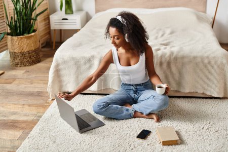 Une afro-américaine frisée dans un débardeur assis sur le sol, entièrement immergée dans son ordinateur portable dans une chambre moderne.