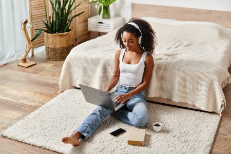 Eine Frau mit lockigem Haar sitzt mit einem Laptop in einem modernen Schlafzimmer auf dem Boden.