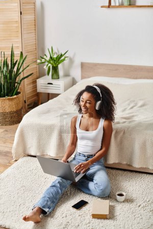 An African American woman with curly hair sits on the floor in a modern bedroom, using a laptop.