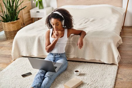 A curly African American woman in a tank top sits on the floor using a laptop in a modern bedroom setting.