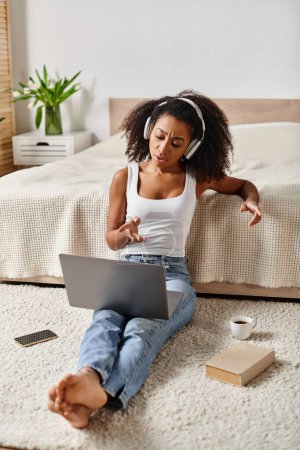 Eine lockige Afroamerikanerin in einem Tank-Top sitzt mit einem Laptop in einem modernen Schlafzimmer auf dem Boden.