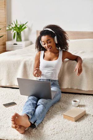 Eine lockige Afroamerikanerin, die in einem modernen Schlafzimmer auf dem Boden sitzt und sich darin vertieft, einen Laptop zu benutzen.