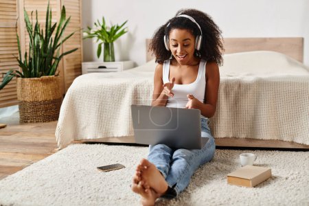 Foto de Una mujer afroamericana en una camiseta sin mangas se sienta en el suelo, centrada en su computadora portátil en un entorno de dormitorio moderno. - Imagen libre de derechos