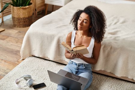A curly African American woman in a tank top is sitting on the floor in a modern bedroom, engrossed in reading a book.