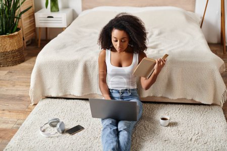 Foto de Mujer afroamericana rizada en camiseta sin mangas sentada en el suelo, enfocada en su computadora portátil en un dormitorio moderno. - Imagen libre de derechos