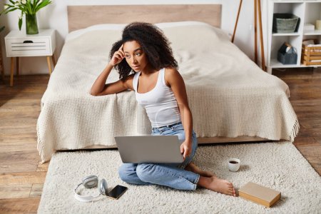 Una mujer afroamericana rizada en una camiseta sin mangas se sienta en el suelo, absorto en el uso de un ordenador portátil en un dormitorio moderno.
