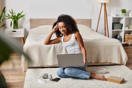 Photo for A curly African American woman in a tank top is sitting on the floor, engrossed in using a laptop in a modern bedroom. - Royalty Free Image