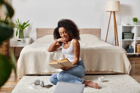 Femme afro-américaine bouclée dans un débardeur assis sur le sol absorbé par la lecture d'un livre captivant dans une chambre moderne.