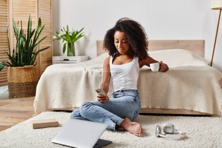 Photo for Curly African American woman sitting on floor next to laptop in modern bedroom. - Royalty Free Image