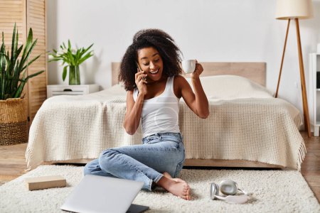 Mujer afroamericana rizada en una camiseta sin mangas sentada en el suelo, absorta en una conversación telefónica en un dormitorio moderno.