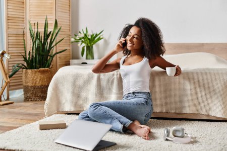 Téléchargez les photos : A curly African American woman in a tank top sitting on the floor, engaged in a phone conversation in a modern bedroom. - en image libre de droit