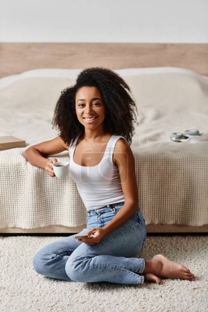 Foto de Mujer afroamericana rizada en una camiseta sin mangas sentada en el suelo frente a una cama en un dormitorio moderno. - Imagen libre de derechos