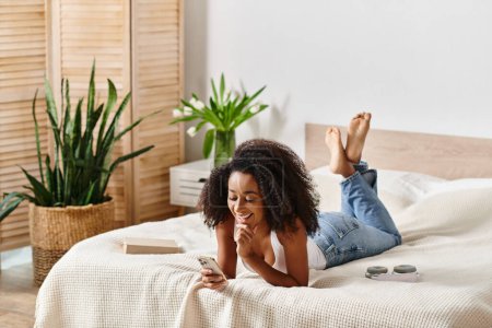 A curly African American woman in a tank top is laying on a bed, absorbed by her cell phone screen in a modern bedroom.