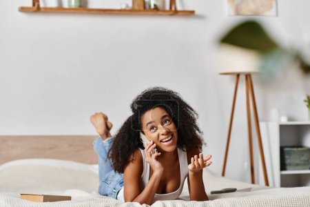 Foto de Una mujer afroamericana rizada en una camiseta sin mangas está cómodamente acostada en una cama, entablando una conversación telefónica en un dormitorio moderno. - Imagen libre de derechos
