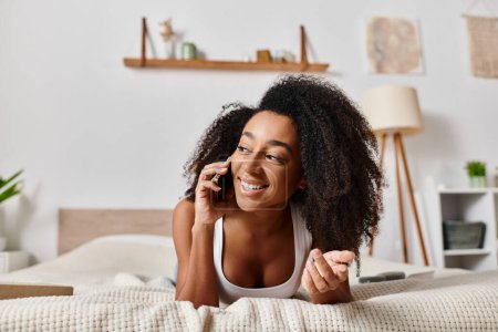 Mujer afroamericana rizada en una camiseta sin mangas tumbada en una cama, absorta en una conversación telefónica.