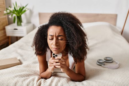 Téléchargez les photos : Femme afro-américaine bouclée dans un débardeur, allongée sur un lit tenant un téléphone portable dans une chambre moderne. - en image libre de droit