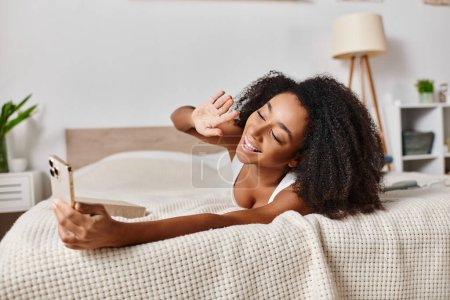 Mujer afroamericana rizada en camiseta sin mangas acostada en la cama, absorbida por la pantalla del teléfono celular en el dormitorio moderno.