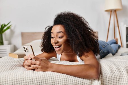 Téléchargez les photos : Une afro-américaine frisée dans un débardeur relaxant sur un lit avec téléphone - en image libre de droit