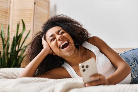 Photo for Curly African American woman in a tank top relaxing on a bed, holding a smartphone in a modern bedroom. - Royalty Free Image