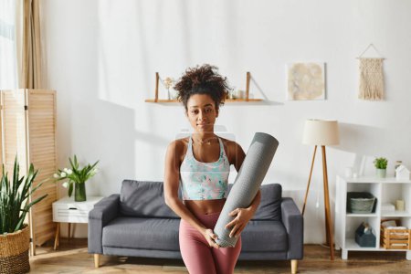 Eine lockige Afroamerikanerin in Aktivkleidung hält eine Yogamatte in ihrem ruhigen Wohnzimmer.
