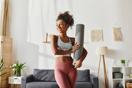 Foto de Una mujer afroamericana rizada en ropa activa sostiene una esterilla de yoga en una elegante sala de estar, lista para su rutina de ejercicios. - Imagen libre de derechos