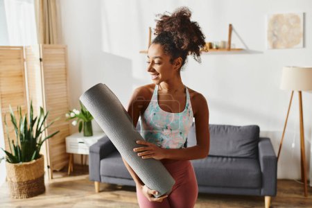 Foto de Mujer afroamericana rizada en ropa deportiva sostiene una esterilla de yoga en una elegante y elegante sala de estar. - Imagen libre de derechos