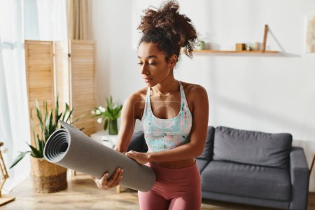 Curly African American woman in active wear stands in a living room, grasping a vibrant yoga mat.