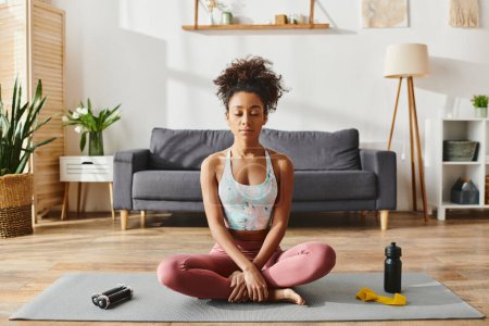 Curly African American woman in activewear practicing yoga on a mat in a cozy living room setting.