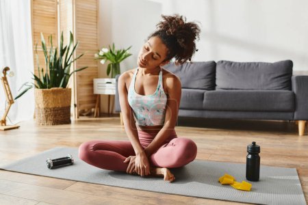 Curly African American woman in active wear sits on yoga mat, practicing mindfulness in cozy living room setting.