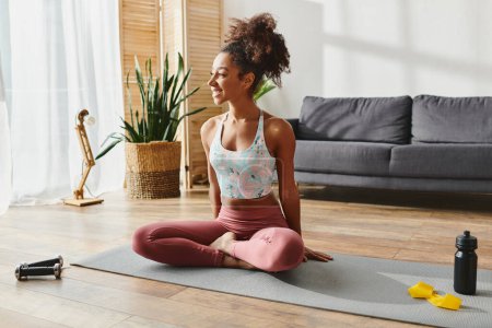 A curly African American woman in activewear serenely practices yoga on a mat in a cozy living room setting.