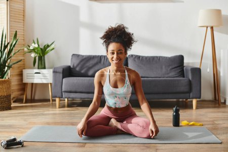 Eine lockige Afroamerikanerin in aktiver Kleidung praktiziert Yoga auf einer Matte in ihrem gemütlichen Wohnzimmer.